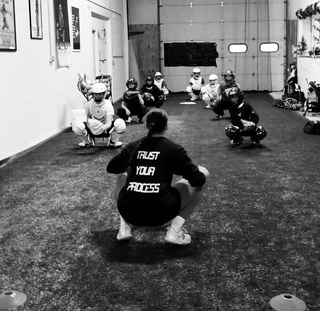 Coach Gabi in front of 8 catchers all in squat position. The back of Coach Gabi's shirt reads "TRUST YOUR PROCESS."