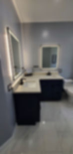 L-shaped double vanity with dark cabinets, white countertops, black faucets, and two illuminated mirrors.