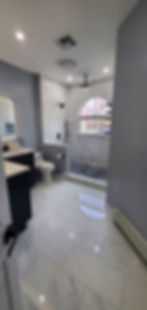 Bathroom overview showing dark vanities, illuminated mirror, white tile flooring, and a custom shower beneath an arched window.
