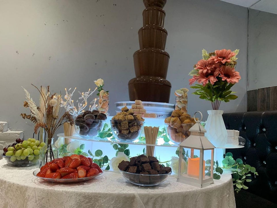 A cascading chocolate fountain overflowing with rich, melted chocolate sits on a table at Zaiqa London restaurant. The table is surrounded by a variety of colorful desserts, including pastries, fruits, and confections.