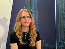 Event interview cover image showing a woman wearing glasses and a microphone, seated against a technical industrial display background