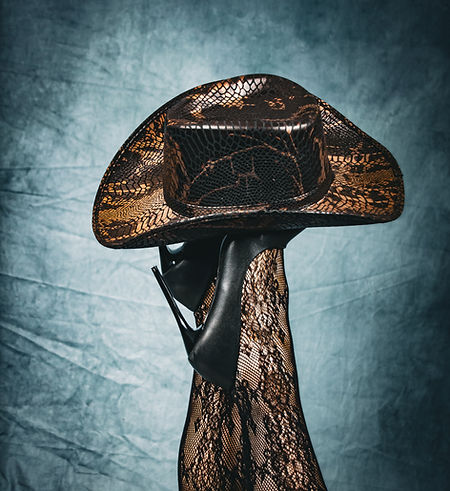 Jambes levées en collants de dentelle noire et escarpins noirs, surmontées d’un chapeau de cowboy aux motifs reptile cuivrés,