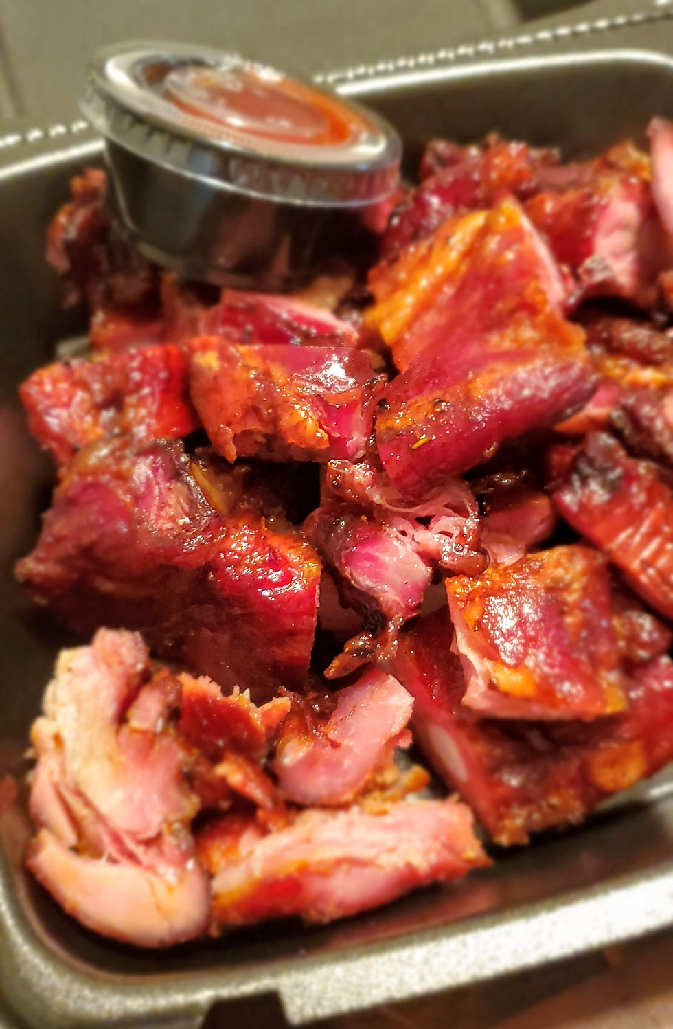 Close-up of juicy, glazed smoked Rib Tips in a tray with a small metal cup of sauce, presenting a hearty and savory meal.