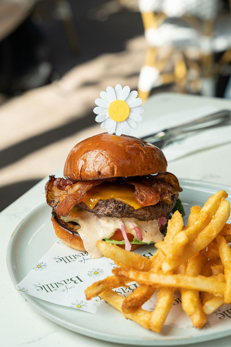 Bacon Cheeseburger on a glossy brioche bun with melted cheddar, pickled onions, tomato and a daisy pick, fries piled alongside