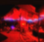 People sit under a red-lit tent with string lights and a drum set, creating a cozy, intimate nighttime gathering