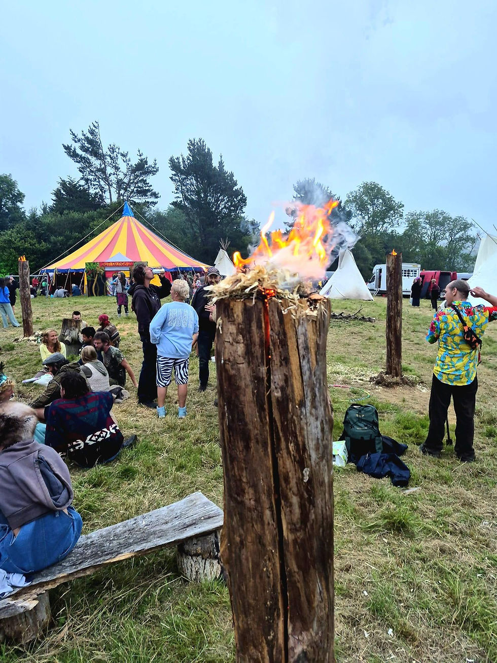 People gather at a grassy festival with teepees and a yellow-pink tent, beside a burning wooden post under a cloudy sky.