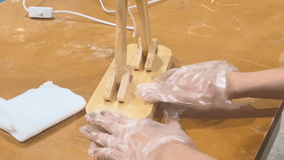 Applying wood wax to a wooden lamp piece with gloves on, ensuring a protective finish.