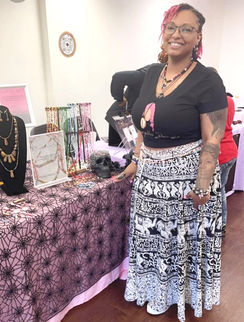 Reecie, wearing a black logo tee and patterned skirt, smiles at her Scorpielle Designs market booth. The display features necklaces, waist beads, and bracelets on a pink spiderweb-themed tablecloth.