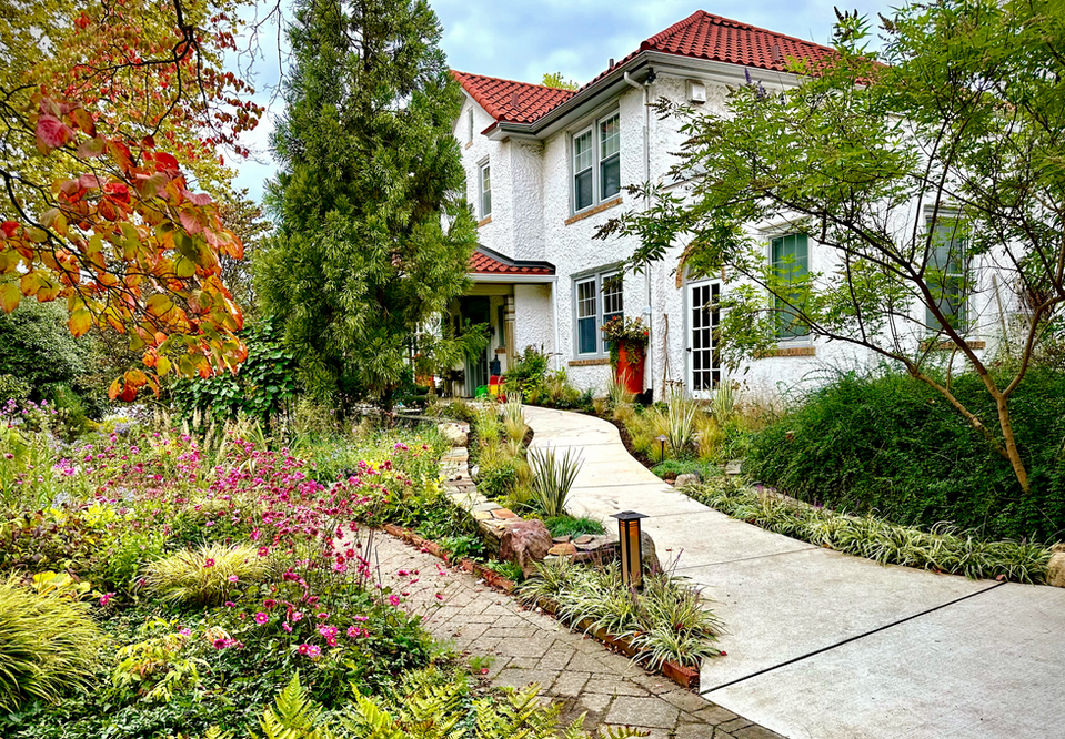 Curving front walk through a lush plant-forward garden beside a white stucco house with red tile roof. a white stucco house with red tile roof.