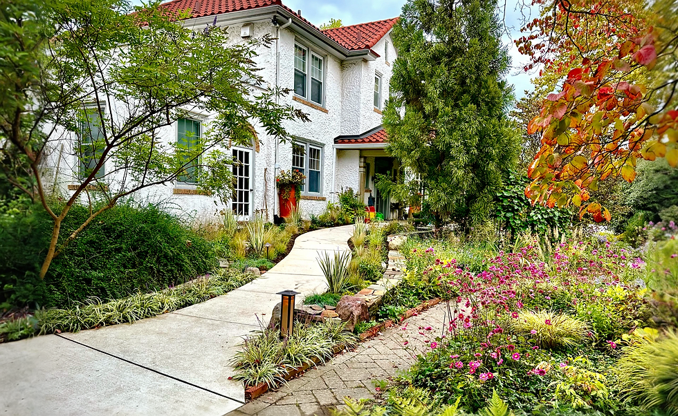 Curving front walk through a lush plant-forward garden beside a white stucco house with red tile roof.