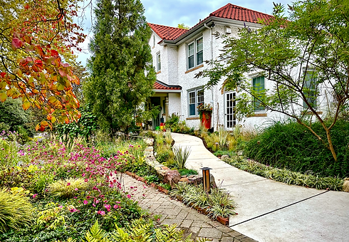 Curving front walk through a lush plant-forward garden beside a white stucco house with red tile roof.