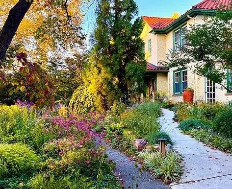 Mediterranean-inspired front garden with curved walkway, white stucco house, red tile roof, layered planting, ornamental grasses, ferns, stonework, and late-season flowers.