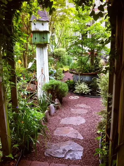 View through a garden arbor toward the house, with stone path, gravel surfacing, layered habitat planting, containers, and birdhouse.