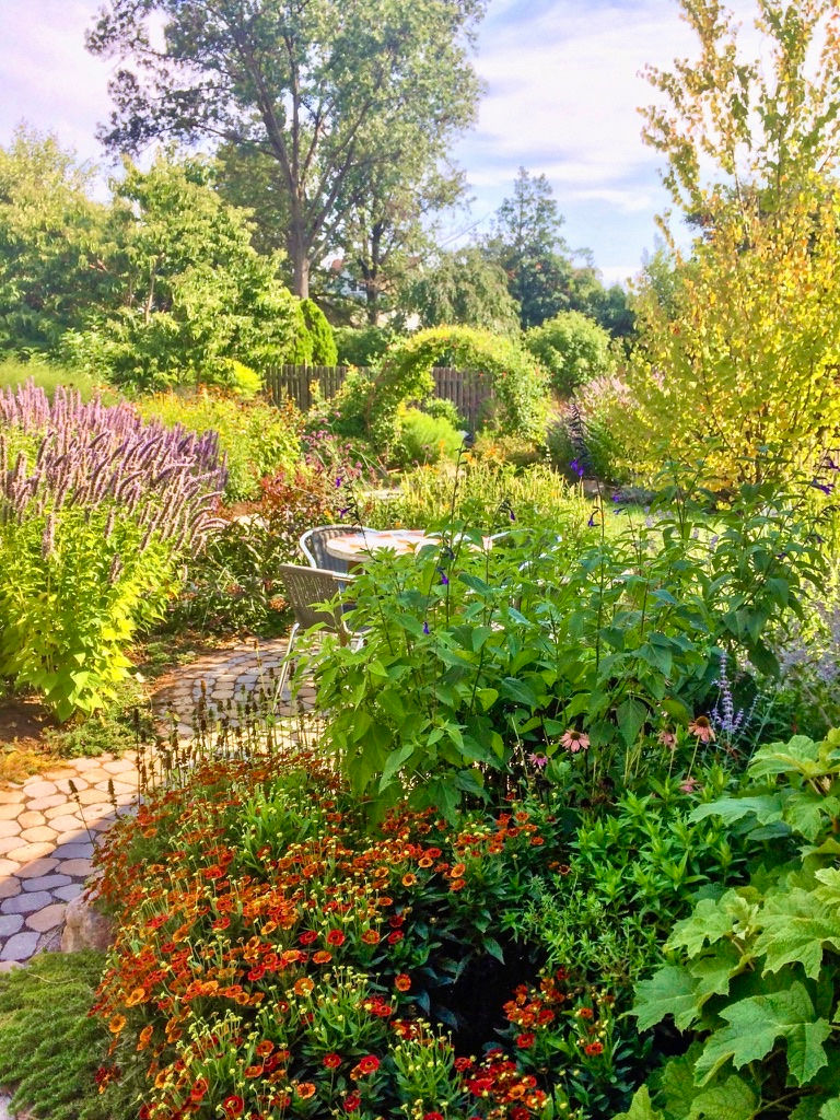 Circular paver sitting area surrounded by lush planting with a vine-covered moon gate in the background.