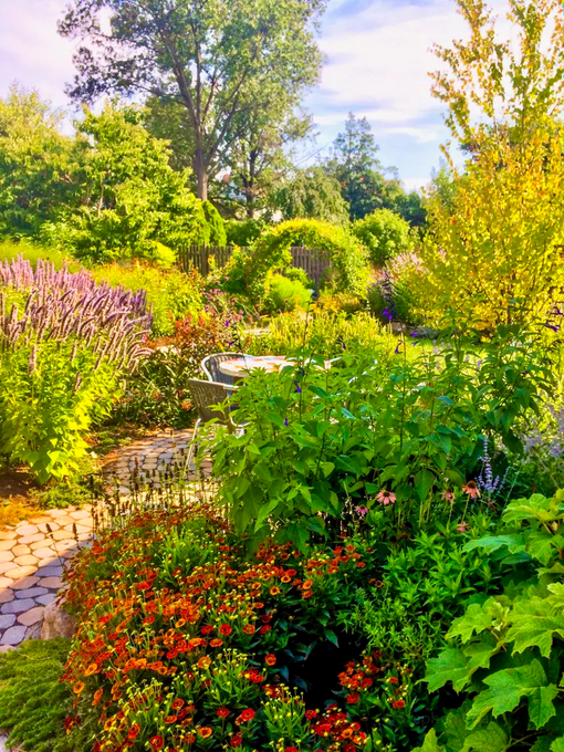 Layered backyard garden with flowering perennials, stone paving, circular seating area, and custom moongate in the distance.