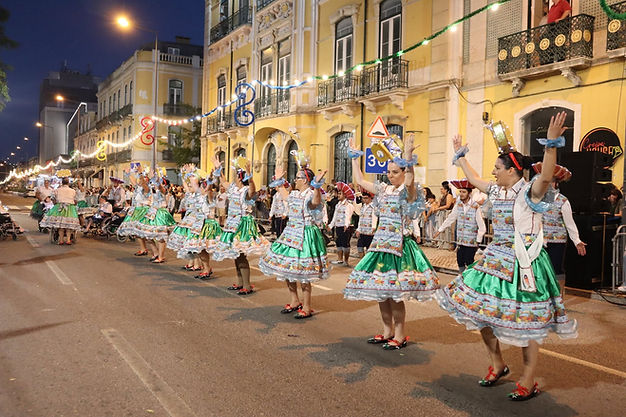 Fotografia noturna de um desfile das Marchas Populares numa avenida iluminada. Alinhadas na estrada, várias marchantes dançam com os braços levantados. Vestem saias verdes brilhantes com folhos e coletes coloridos estampados com desenhos de peixes. Ao fundo, veem-se edifícios antigos de cor amarela e público a assistir atrás de vedações.