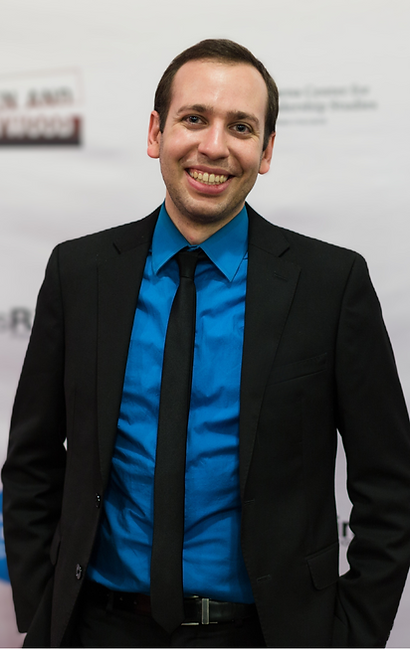 Headshot: Avi stands in a suit in front of a step-and-repeat