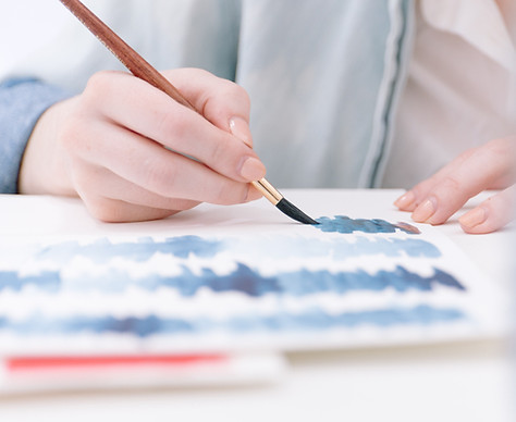 A person's hand painting blue wiggly lines with watercolours on white paper..