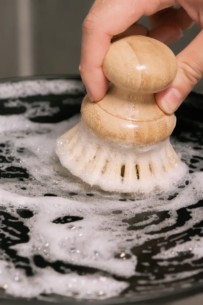 Thumbnail: Hand scrubbing dark pan with a wooden dish brush and soap suds.