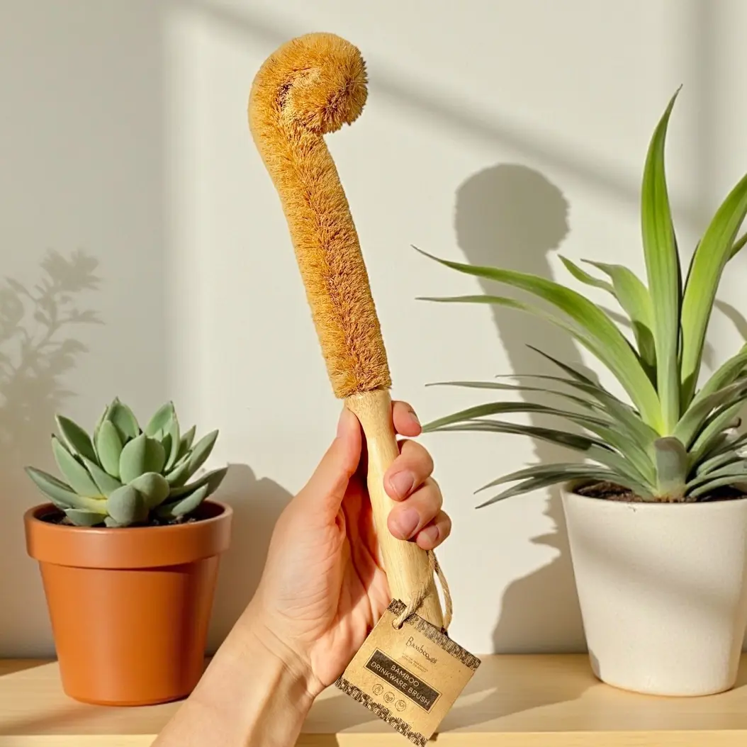 Person's hand holds natural fiber bottle cleaning brush, plants in background.