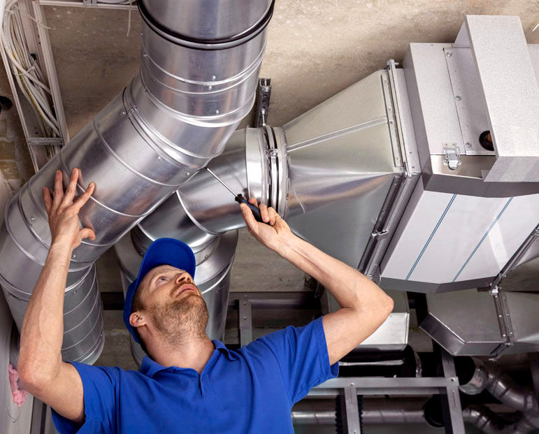 A worker in a blue uniform and cap adjusts ducts on a ceiling with a screwdriver. The setting is industrial with silver metal pipes.