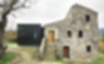 Stone house with wooden doors next to a modern black extension. Leafless tree in the foreground, overcast sky in rural setting.