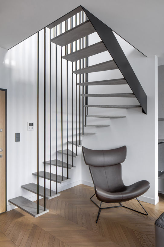 Hanna Daniłów - Penthouse dla minimalisty, Warszawa, 2023 r. Widok strefy dziennej. "Lewitująca" klatka schodowa, ze stopniami mocowanymi wspornikowo do ścian oraz stalowego płąskownika, po środku zawieszone na stalowych prętach. 