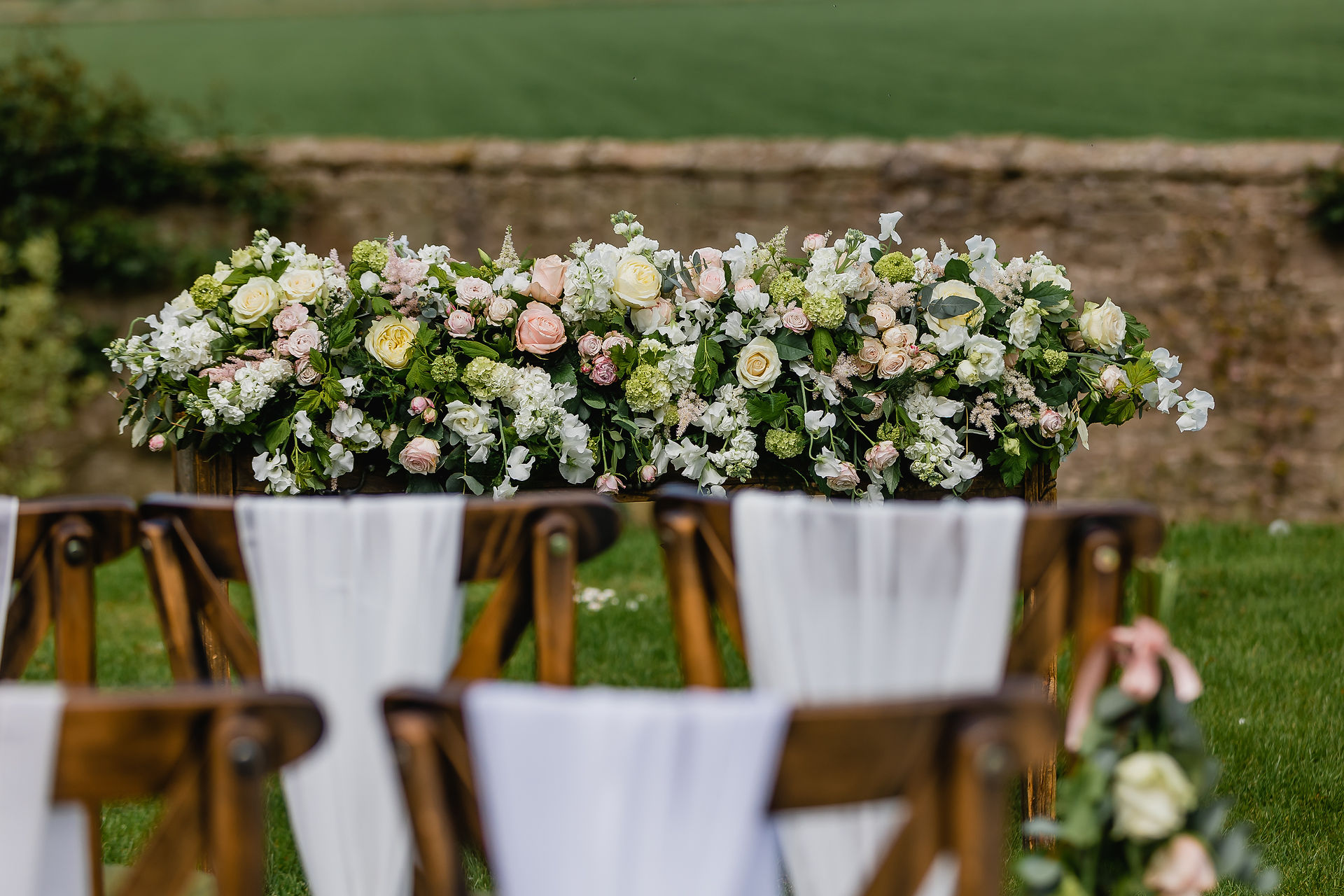 Wedding ceremony at Newton Hall's Wedding Hill, in the beautiful Northumberland countryside. An outdoor ceremony a full length floral display on the registrar table, flowers on the aisle chairs, white chiffon chair drops