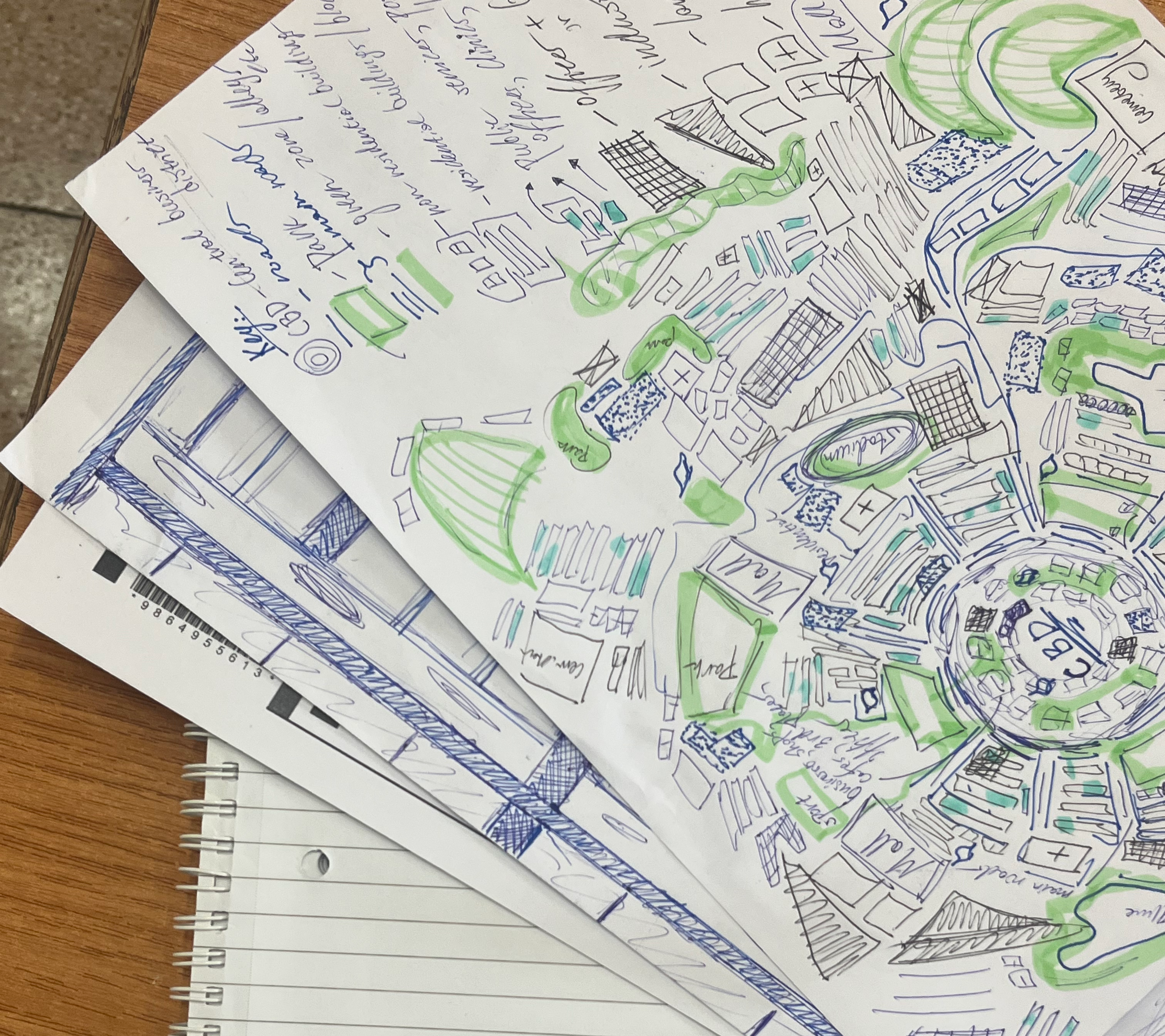 Stacked sketch papers and a spiral notebook on a desk, covered with blue and green doodles and notes