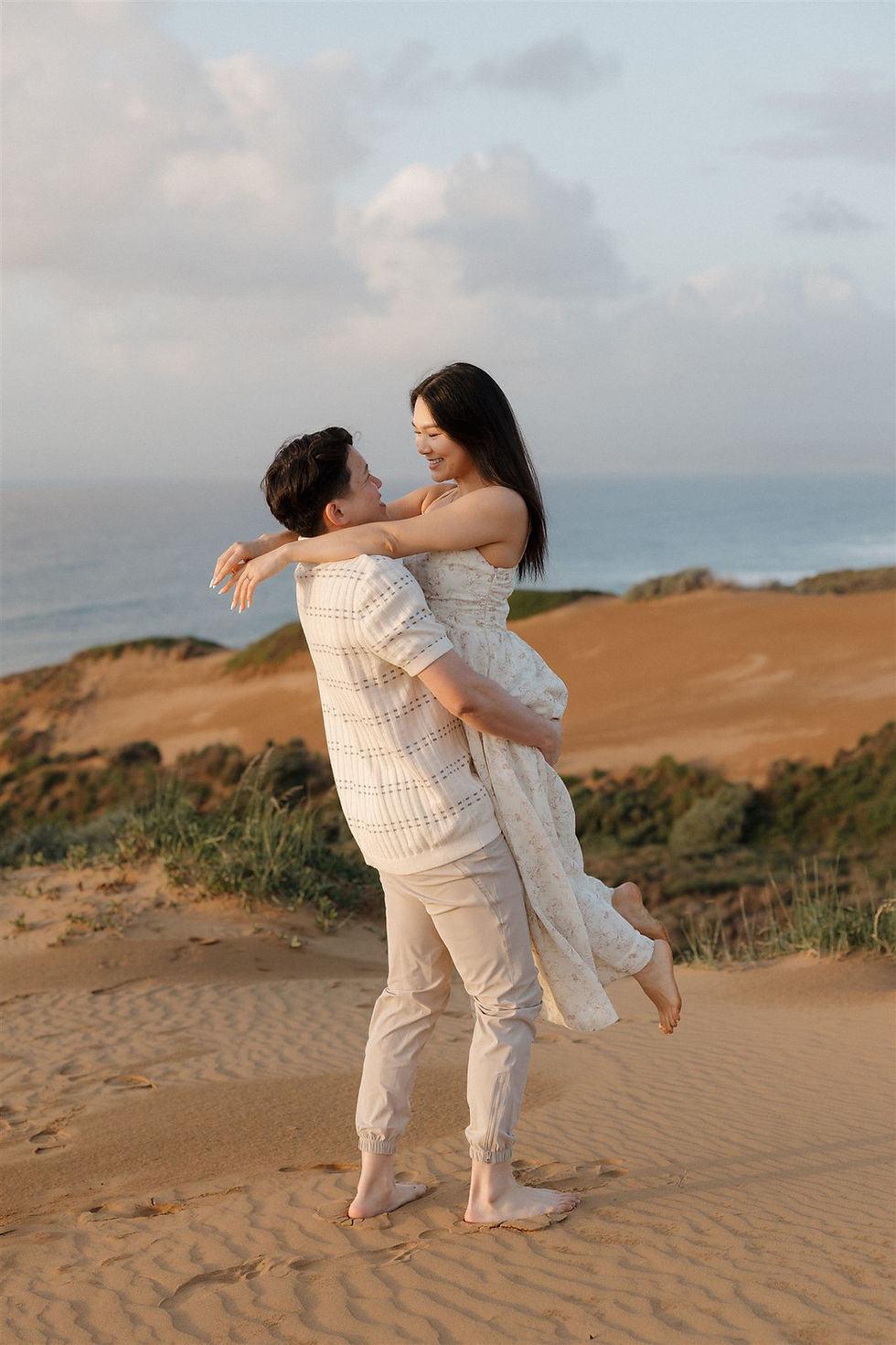 A man lifts a smiling woman on a sandy beach with ocean and hills in the background. They wear light clothing, creating a serene, joyful scene during Central Coast Proposal photographed by Mackenzie Rana 