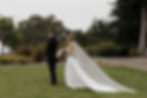 Bride in a white gown holds flowers, standing behind groom in black suit. They're outside on grass, surrounded by trees and shrubs at Cypress Ridge capture by a San Luis Obispo Photographer Mackenzie Rana