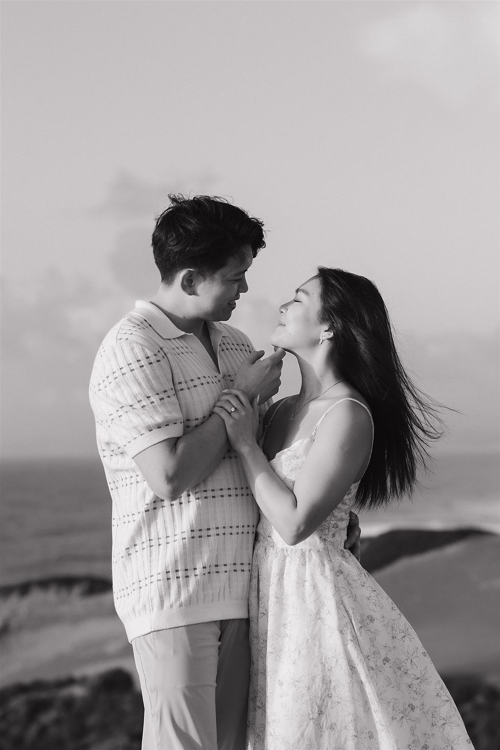 A couple embraces on a windy day atop a scenic hillside, gazing into each other's eyes. Black and white image with an ocean backdrop by Proposal Photographer on the Central Coast Mackenzie Rana Photography