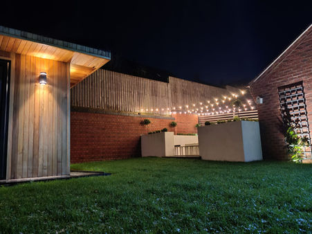 Image shows a green lawn, brick house and wooden outhouse building at night illuminated by lights installed by Jet Electrical.