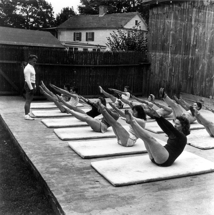 Women exercise Pilates outdoors on mats, lifting legs in unison, while an instructor stands by. Wooden fence and house in the background.