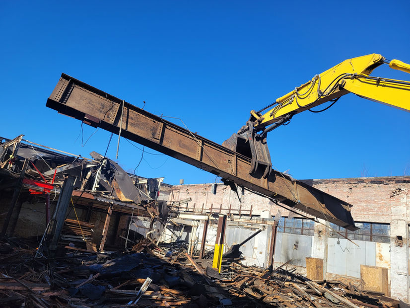 The arm of an excavator holds up a large steel, beam over a pile of rubble during the demolition of a building.