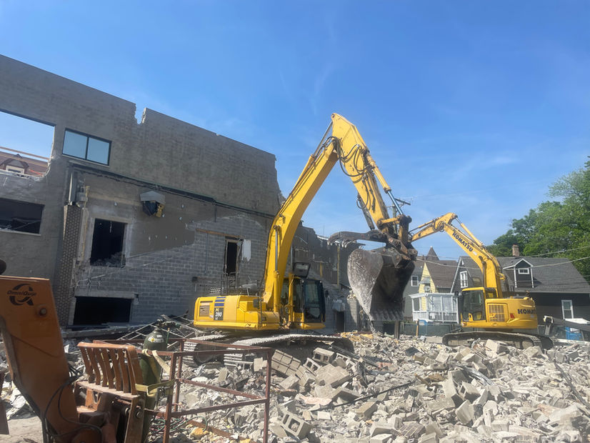 On a bright day, two excavators demolish a building while placing rubble, cement bricks, and garbage in appropriate piles.