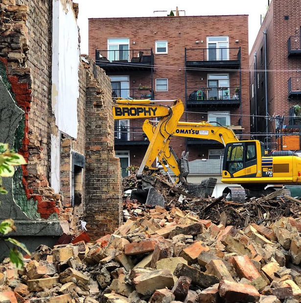 Two excavators (one behind a wall) demolish a building while surrounded by apartments with piles of bricks laying on the ground.