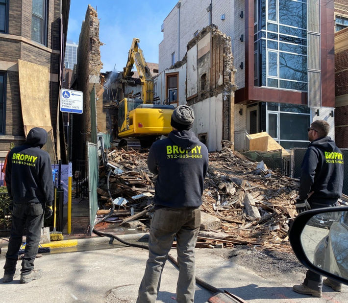 Three male demolition specialists stand to watch one of Brophy Excavation's excavators safely demolish a building without harming the neighboring buildings.