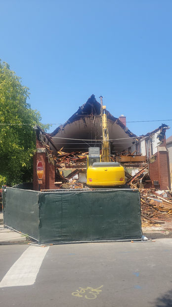 On a sunny day, a yellow excavator tears down a building. Protective fences surround the building for the public's safety against rubble. Various trees can be seen on the left.