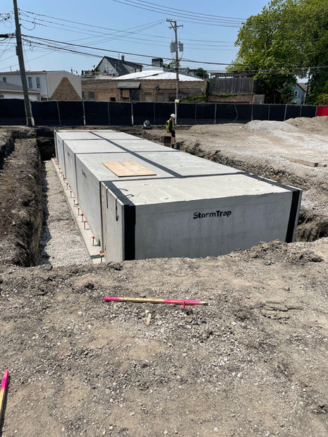 A concrete structure lays in a large, flat hole in the ground. A person in protective personal equipment walks around and ensures proper installation.