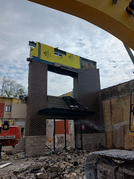 Two machines demolish a jobsite as a hose splashes water on the area to minimize dust and irritants in the air. Parts of a building still standing can be seen.