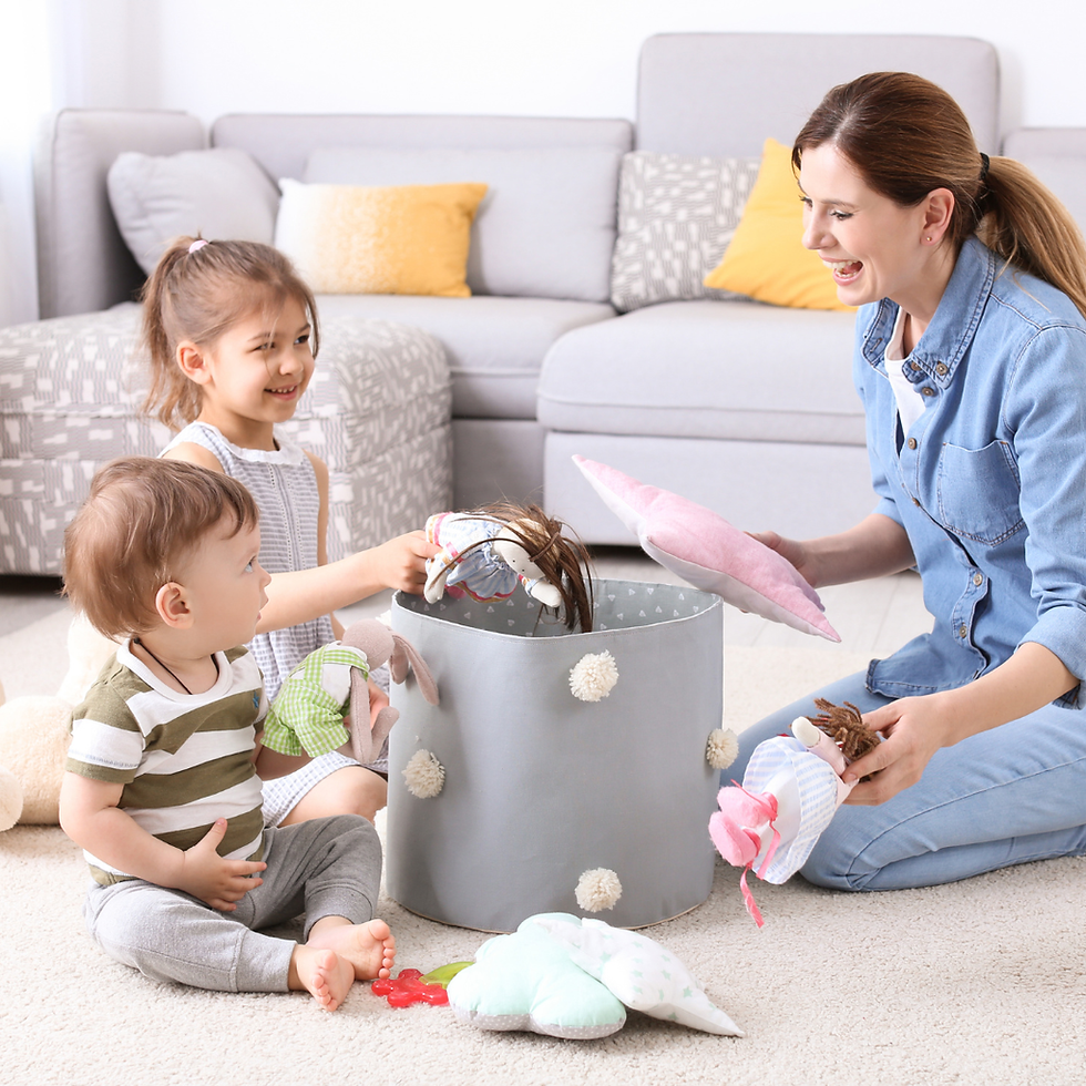Smiling mother and two children tidying dolls and tosy into a gray toy bin in a bright living room with a sofa and pillows.