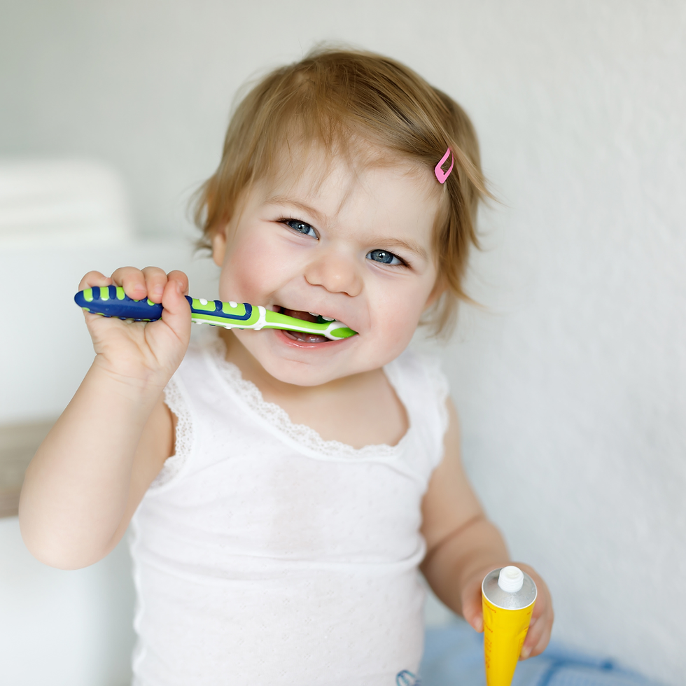 Smiling toddler in a white top brushing teeth with a green-and-blue toothbrush, holding a yellow toothpaste tube against a soft white background