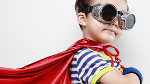 Boy in silver goggles and a red cape, wearing a striped shirt, posing like a superhero against a white background.