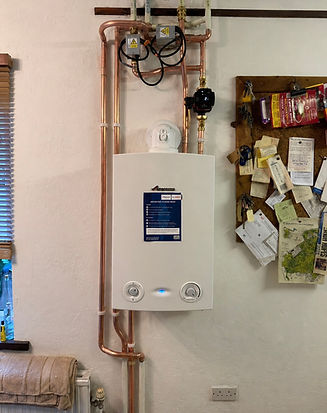 Wall-mounted white boiler with copper pipes and a decorative wreath on a light-colored wall