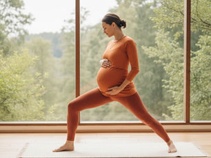 Hamile bir kadın turuncu yoga kıyafetiyle, cam kenarında doğa manzarasına karşı poz veriyor. Huzurlu ve odaklanmış görünüyor.