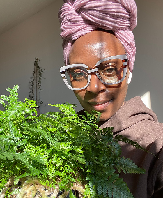 Black woman with pink headwrap, wearing pink and brown framed glasses, in a brown hoodie, holding a rabbit's foot fern houseplant