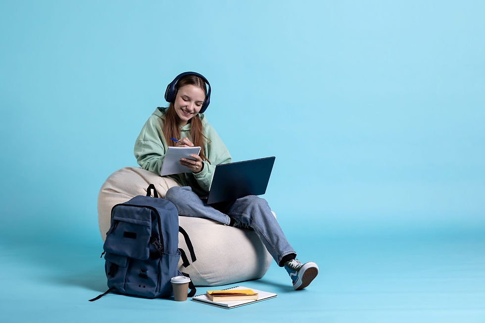 Smiling student wearing headphones studying with laptop and notepad, sitting on bean bag chair with backpack and books on blue background.