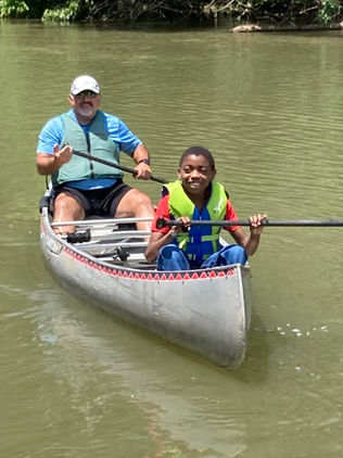 a man sits in the rear of a floating canoe with a young boy in the front. Both holding paddles with smiles. 