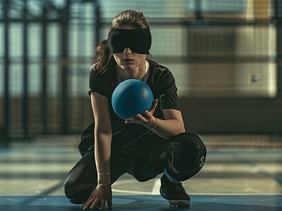 a crouched goalball player wearing an eye mask with one hand on the floor and the other holding a goalball